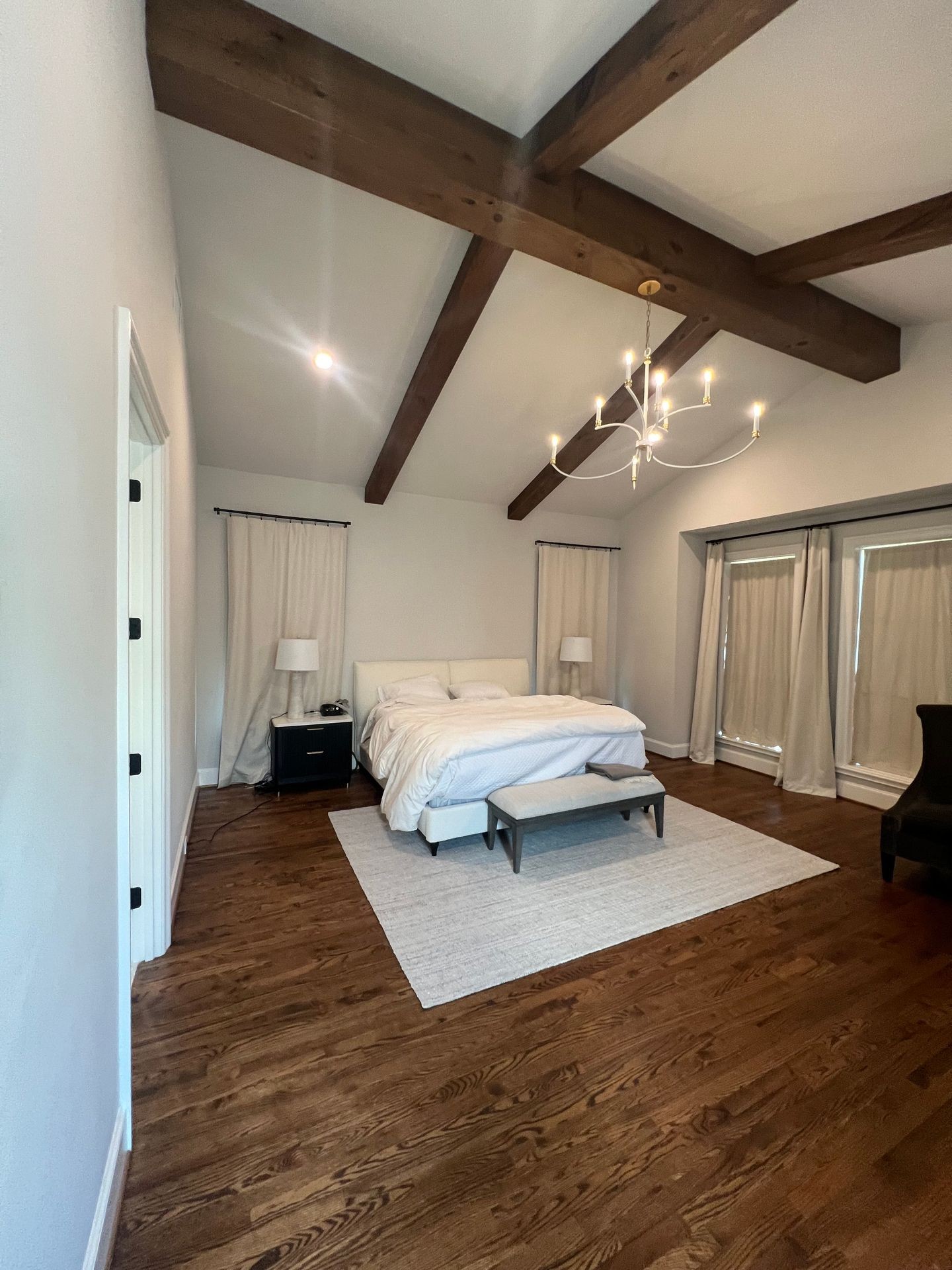 Cozy bedroom with wooden beams, modern chandelier, white bed, and dark wood flooring.